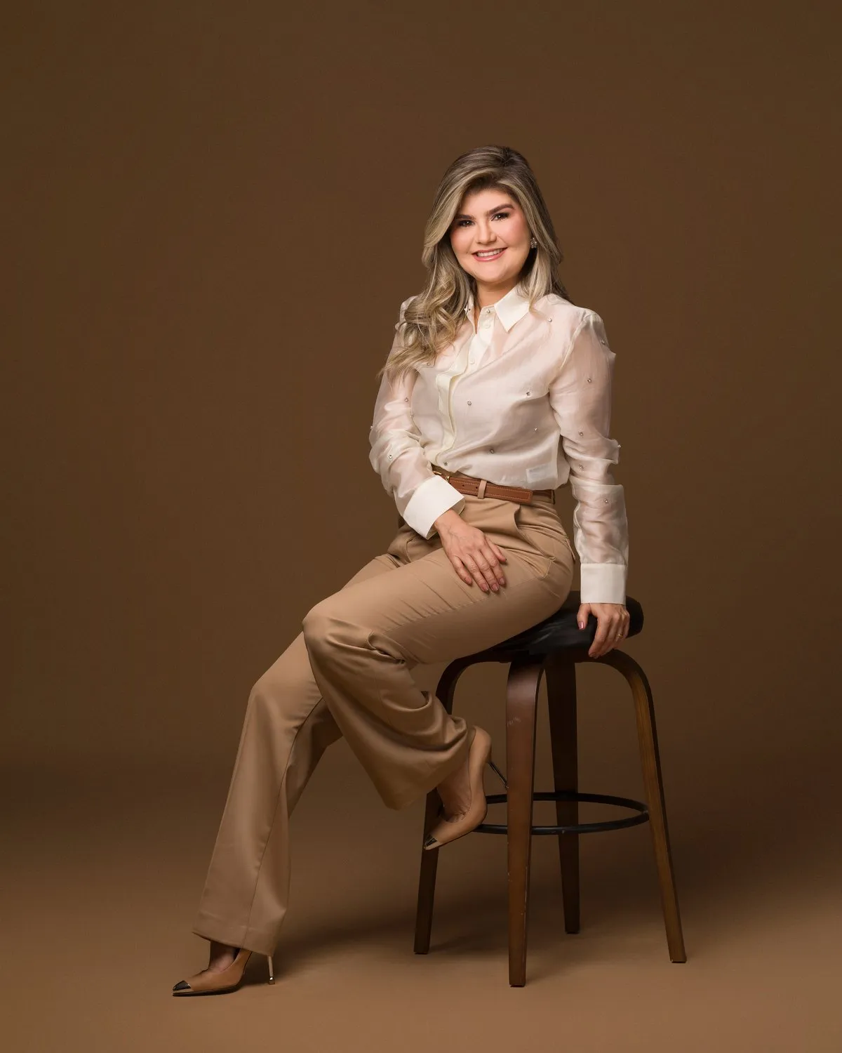 Raquel Dias sentada em uma banqueta, de camisa clara e calça bege