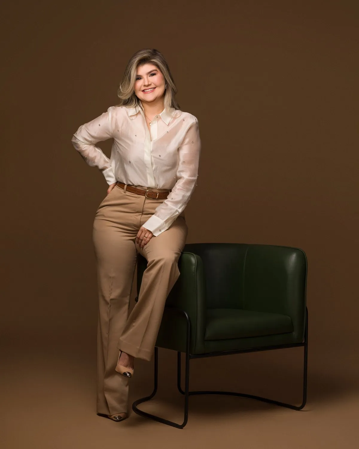 Raquel Dias de pé, com a mão na cintura, ao lado de uma poltrona verde
