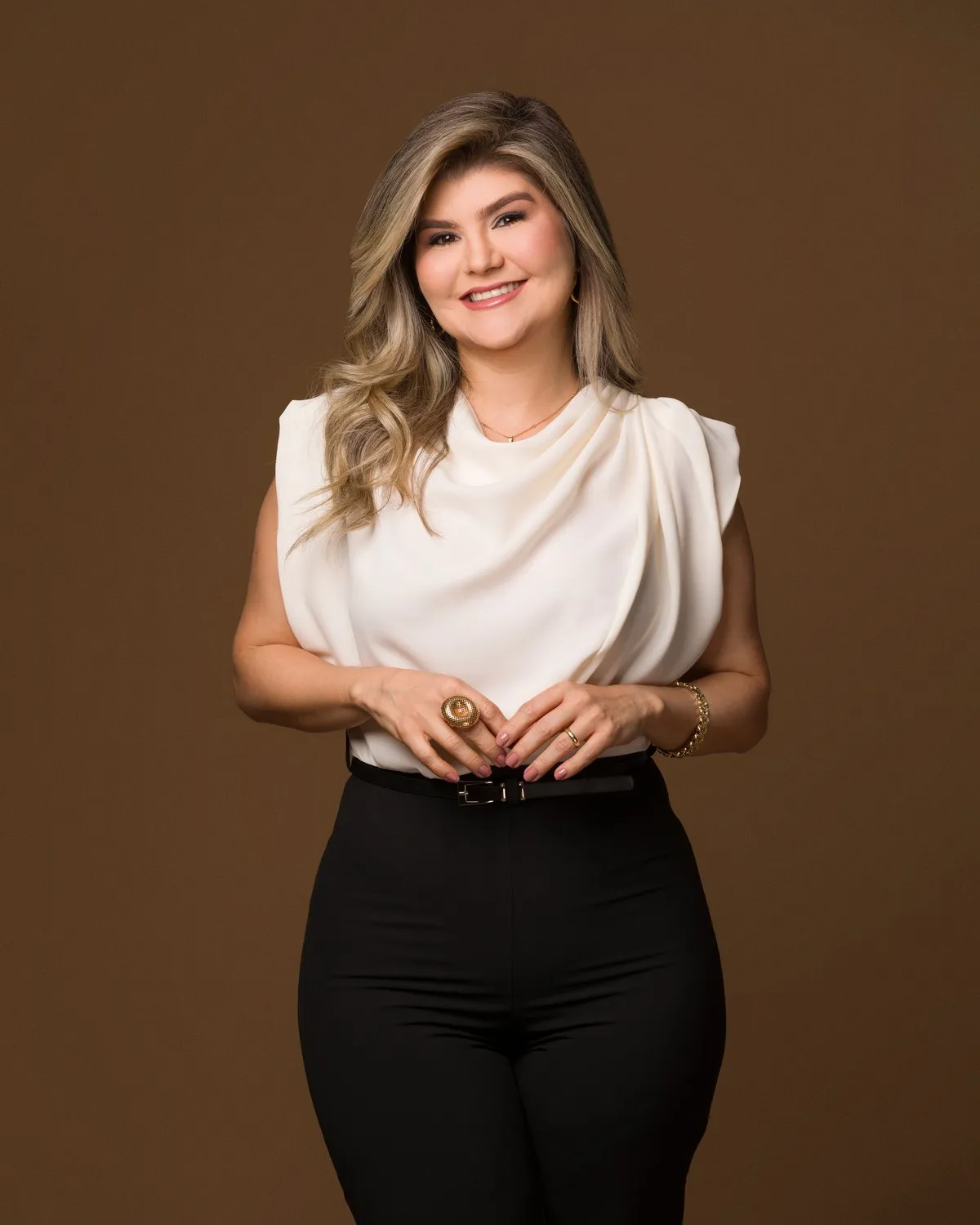 Raquel Dias de pé, de blusa clara e calça preta, em fundo marrom